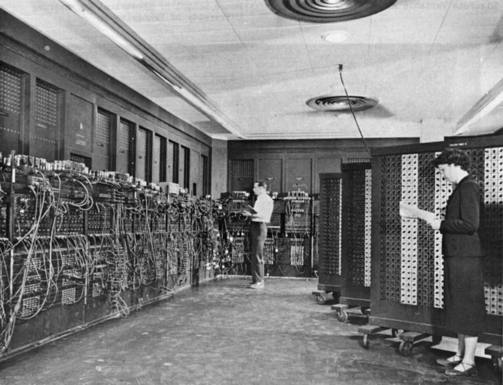 Black-and-white photo of two people working at the ENIAC computer, which fills a room with tall cabinets covered in cables and switches.